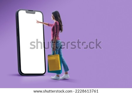 Happy woman doing online shopping using a big smartphone, she is holding shopping bags and touching the blank screen Royalty-Free Stock Photo #2228613761