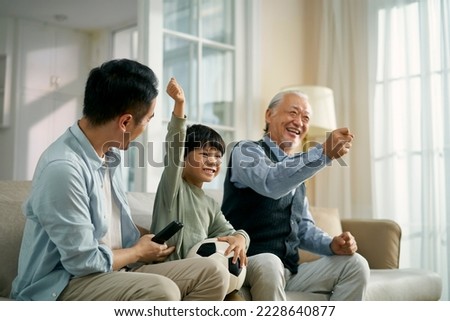 asian son father grandfather sitting on couch at home celebrating goal and victory while watching live broadcasting of football match on TV together Royalty-Free Stock Photo #2228640877