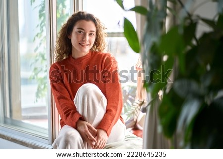 Pretty serene young woman sitting on windowsill relaxing at home looking at camera. Smiling calm lady chilling in apartment, dreaming, thinking of peaceful time enjoying peace of mind. Portrait Royalty-Free Stock Photo #2228645235