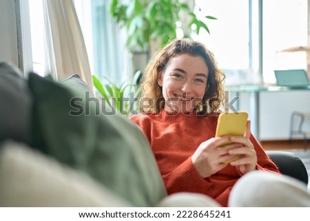 Smiling young pretty woman sitting on couch using apps on cell phone technology, happy lady holding smartphone in hands, looking at camera, relaxing on sofa with cellphone checking cellular device.