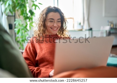Relaxed happy young woman sitting on sofa using laptop at home surfing, doing online ecommerce shopping, looking at computer ordering sale products on website, watching videos or elearning. Royalty-Free Stock Photo #2228645257