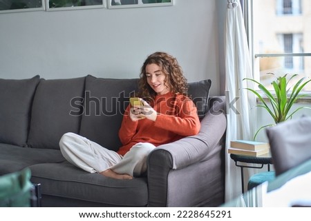 Young woman sitting on couch holding smartphone, looking at cellphone using cell phone checking mobile apps, reading messages, doing ecommerce shopping, chatting online, watching videos, playing game.