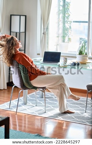 Young smiling pretty woman relaxing at home sitting on chair at table taking break after computer remote work or study. Happy calm lady feeling pleasure working from home. Vertical
