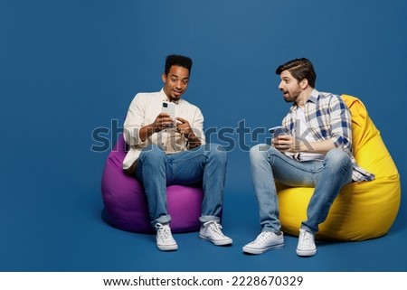 Full body young two friends shocked men wear white casual shirts together sit in bag chair hold in hand use show mobile cell phone isolated plain dark royal blue background. People lifestyle concept Royalty-Free Stock Photo #2228670329