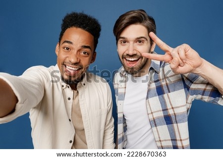 Close up young two friends men wear white casual shirts together do selfie shot pov on mobile cell phone cover eye with v-sign isolated plain dark royal navy blue background. People lifestyle concept