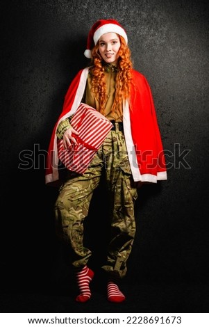 A red-haired woman in a red hat holds a gift box in her hands. Portrait of a happy girl in a military army uniform on a black dark background. The concept of Christmas and New Year.