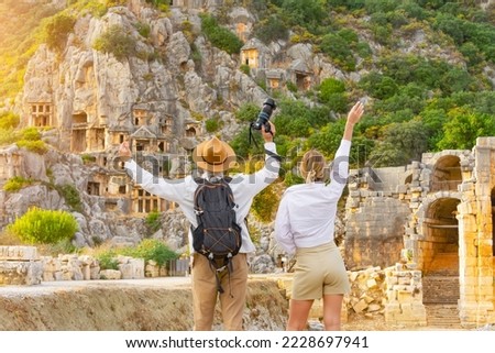 Guy with backpack in hat girl, travelers raising their hands up in surprise, look at ruins ancient city, ancient unique famous places of Turkey, Antalya Demre, Mira, ancient Lycian tomb casts faces Royalty-Free Stock Photo #2228697941