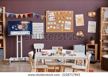 Interior of spacious classroom for nursery schoolkids with table surrounded by chairs in center and boards with notes and pictures
