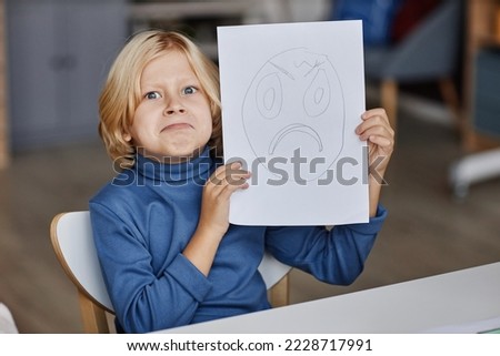 Cute blond boy imitating expression of gloomy face drawn on paper that he showing to you while sitting by table at lesson of drawing Royalty-Free Stock Photo #2228717991