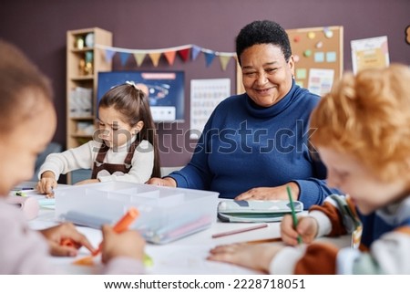 Smiling mature teacher of nursery school looking at one of learners drawing with crayons while sitting among intercultural kids Royalty-Free Stock Photo #2228718051