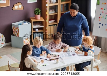 Group of adorable learners of nursery school sitting by table at lesson while mature African American female teacher standing by them