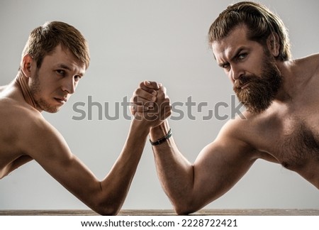 Arm wrestling. Heavily muscled bearded man arm wrestling a puny weak man. Arms wrestling thin hand, big strong arm in studio. Two man's hands clasped arm wrestling, strong and weak, unequal match.