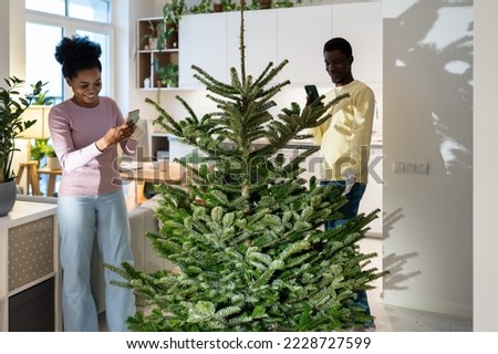 Young African woman taking photo of fresh undecorated christmas tree for sharing in social media. Happy couple busy install first xmas pine in new home prepare for new year winter holidays together