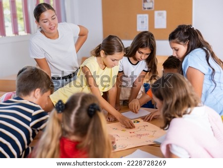 Group of interested school kids with female teacher sitting together around desk in classroom, playing educational tabletop game Royalty-Free Stock Photo #2228728269