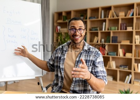 English course. Happy arab male teacher having foreign language class sitting near whiteboard with grammar rules, talking and smiling to camera. Education concept