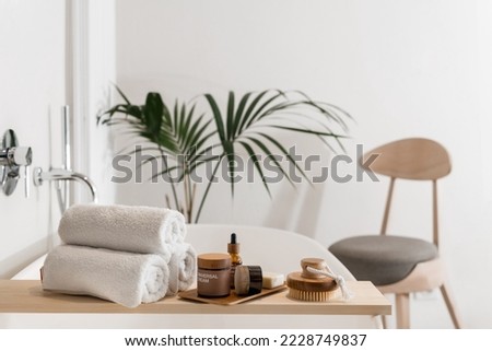 skin care cosmetics on bamboo tray, massage peeling brush and fresh terry towels folded on wooden caddy on bathtub, plant and chair in apartment with modern interior Royalty-Free Stock Photo #2228749837