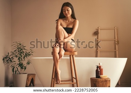 Asian girl going to take morning bath, sitting on high wooden stool near bathtub, enjoying day off, looking peaceful and happy