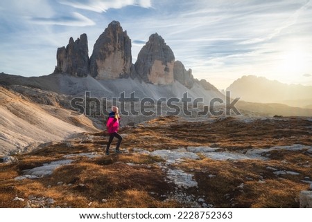 Walking girl with backpack on the trail in mountains at sunset in autumn. Tre Cime, Dolomites, Italy. Beautiful landscape with young woman, high rocks, path, stones, orange grass, sky in fall. Hiking