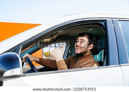 modern satisfied chauffeur bearded young man sitting in a car shows thumb up from window. Man smiles, mouth wide open. Portrait of happy man showing ok sign