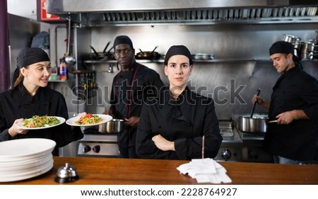 Portrait of focused woman head chef posing in modern restaurant kitchen, team of chefs working behind Royalty-Free Stock Photo #2228764927
