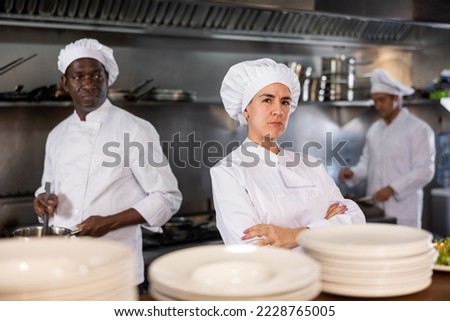 Confident woman executive chef standing in restaurant kitchen, posing on camera, while his team working Royalty-Free Stock Photo #2228765005