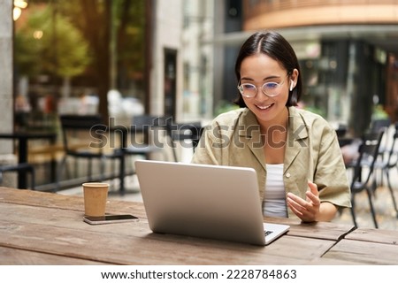 Young woman sitting on online meeting in outdoor cafe, talking to laptop camera, explaining something, drinking coffee. Royalty-Free Stock Photo #2228784863