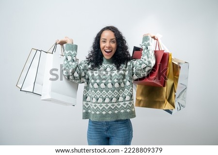 Portrait of smiling Latin woman with curly hair wearing sweater with shopping bag isolated on light blue background.
