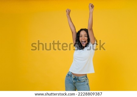 Happy curly Latin woman in white t-shirt raise hands up isolated on yellow background. Royalty-Free Stock Photo #2228809387