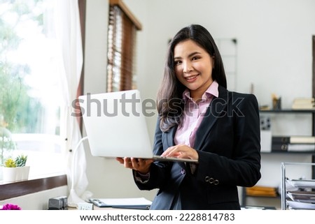 Young businesswoman in suit is standing and holding laptop to typing data while working about analysis of strategy and planning new business in office.