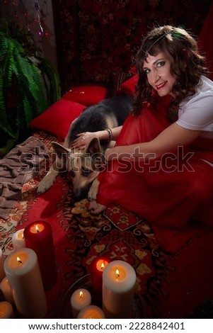 Beautiful arabian girl with candles and big dog in red room full of rich fabrics and carpets in sultan harem. Photo shoot of woman an oriental style odalisque. Model poses as a caring wife and hostess