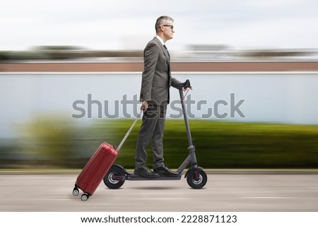 Corporate businessman riding an eco-friendly electric scooter and carrying a roller suitcase, city in the background, sustainable mobility concept