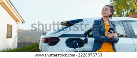 Young woman with smartphone waiting while her electric car charging in home charging station, sustainable and economic transportation concept. Royalty-Free Stock Photo #2228875753