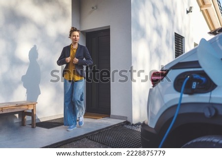 Young woman preparing to work while her electric car charging in home, sustainable and economic transportation concept. Royalty-Free Stock Photo #2228875779