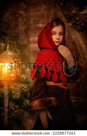 A pretty little girl in medieval clothes stands by a wooden house and burning lantern on a snowy winter night. Wooden house background. Winter's tale. Christmas and New Year concept.