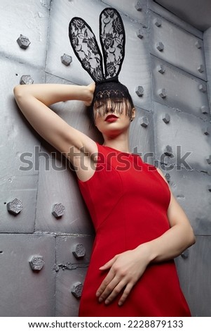 Young woman in black rabbit or hare fancy mask and red dress. Eyes closed. Hands up. In the background there is a metal wall with rivets . Royalty-Free Stock Photo #2228879133