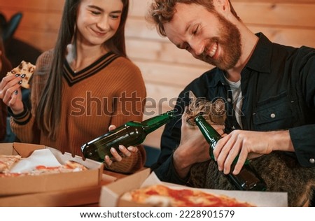 Man and woman sitting indoors and pretending that they giving beer to the cat.