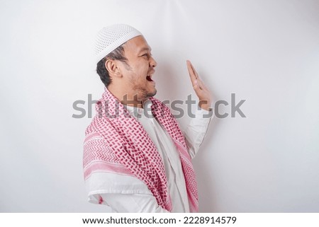 Excited Asian Muslim man pointing at the copy space beside him, isolated by white background