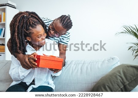 Young couple is at home. They are sitting on a couch, head to head and holding a present. Cheerful young woman receiving a gift from her boyfriend. Shot of a loving husband giving his wife a gift.