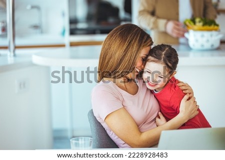 Positive young woman helping daughter in searching information for homework on internet while sitting together at table with laptop at home. Smiling mother and kid having fun