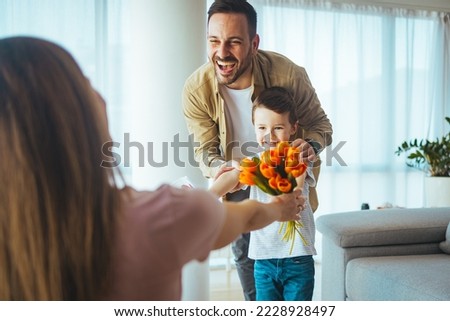 Cheerful mother hugging son and reading handmade greeting card with heart while resting on sofa during holiday celebration mothers day at home. Happy woman receiving flowers and greeting card 