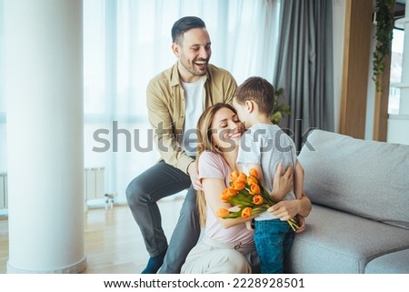 Cheerful little boy with card and with bouquet of tulip flowers smiling and congratulating happy mom on mother day at home. Smiling woman looking at adorable son while sitting on floor 