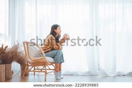 Happy asian woman relaxing drinking hot coffee or tea in holiday morning vacation on armchair at home, Cosy scene, Smiling pretty woman drinking hot tea in autumn winter. copy space. Royalty-Free Stock Photo #2228939885