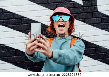 Excited redhead woman screaming while taking a selfie photo outdoors. Emotional hipster fashion women in bright clothes, heart shaped glasses, bucket hat taking selfie photo on the phone camera