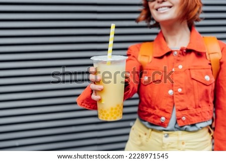 Glass of fruity sugar flavored tapioca pearl bubble tea with straw in hand of no face hipster fashion woman in bright clothes on the gray striped wall background. Selective focus. Copy space.