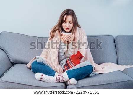 Woman freezes in wintertime. Young girl wearing warm woolen socks and wrapped into two blankets, holding a cup of hot drink and heating pad while sitting on sofa at home. Keep warm. Selective focus. Royalty-Free Stock Photo #2228971551