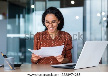 Young happy and successful businesswoman in glasses working with documents inside office, Hispanic woman with laptop looking at bills and contracts, financier with curly hair using laptop.