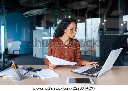 Young happy and successful businesswoman in glasses working with documents inside office, Hispanic woman with laptop looking at bills and contracts, financier with curly hair using laptop. Royalty-Free Stock Photo #2228974299