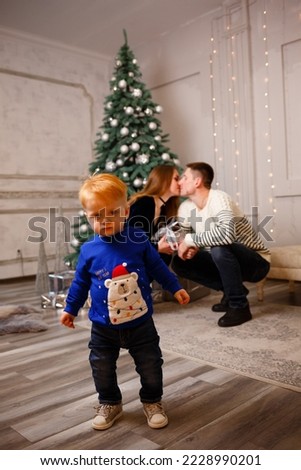 Focus on the little boy. In the background behind him, mom and dad are kissing near the Christmas tree