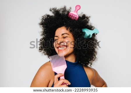 Beautiful woman with Afro hair having fun with fork combs in a studio. Smiling young woman singing and using a comb as a mic. Happy woman of color celebrating her natural hair with pride. Royalty-Free Stock Photo #2228993935