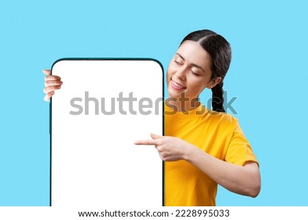 Advertising. A young pretty Caucasian woman smiles and points her finger at mock up screen of a large smartphone. Isolated on blue background. The concept of online shopping and social media.
